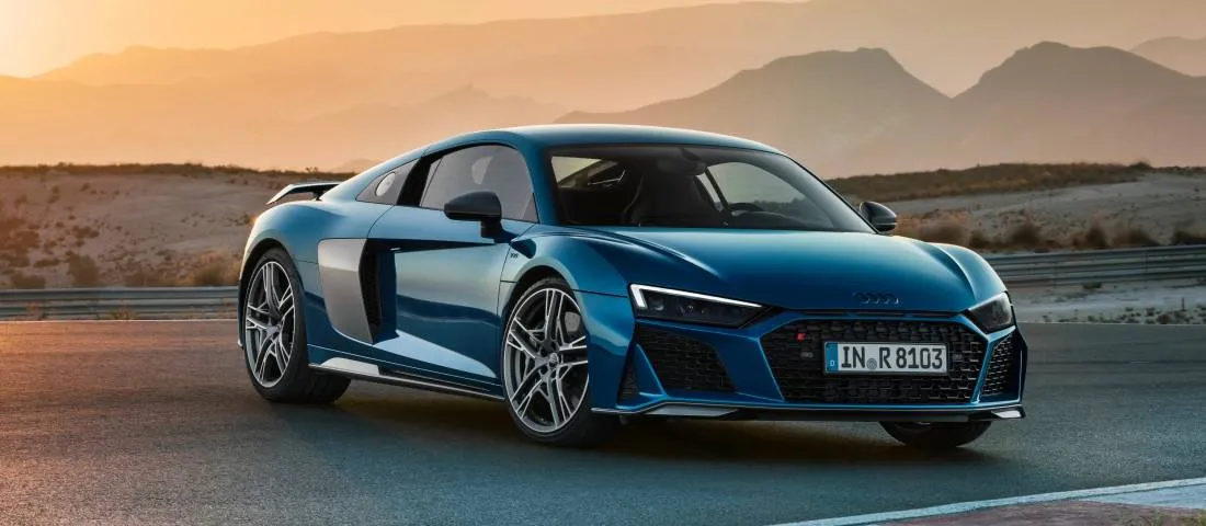 "Audi R8 blu parcheggiata su una strada, con un suggestivo tramonto e montagne sullo sfondo."